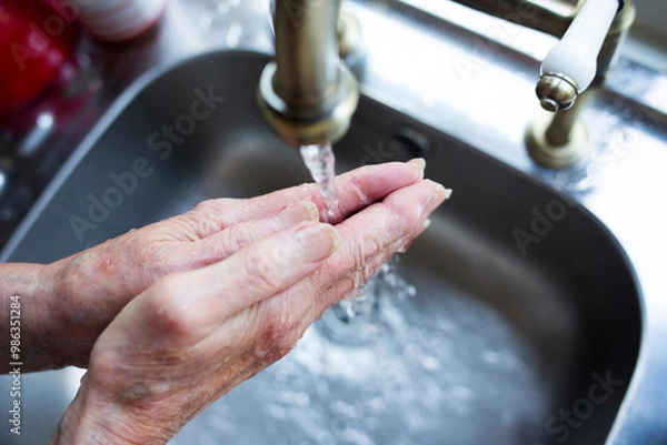 Obraz Close-up of elderly lady washing her hands under a running tap against a silver shade sink 