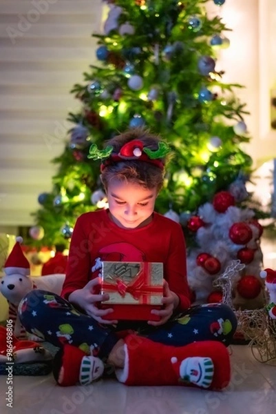 Fototapeta Happy little boy sitting in front of a christmas tree and suprisingly looking at the gift