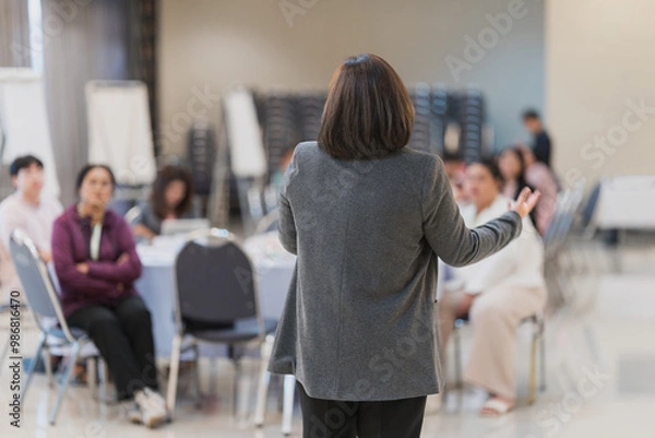 Fototapeta Speaker Leading Business Workshop for Professional Audience. A presenter conducts a business workshop, addressing a group of professionals seated at tables in a modern conference room.