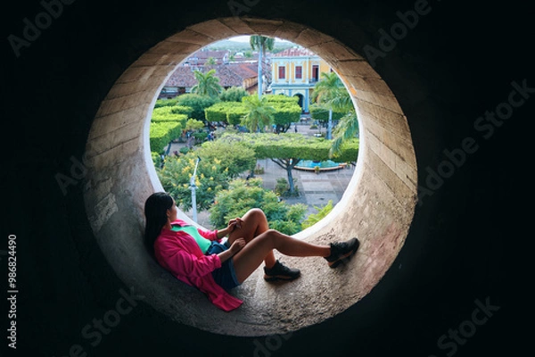 Obraz Woman Relaxing in Circular Window Overlooking Lush Park in Granada, Nicaragua