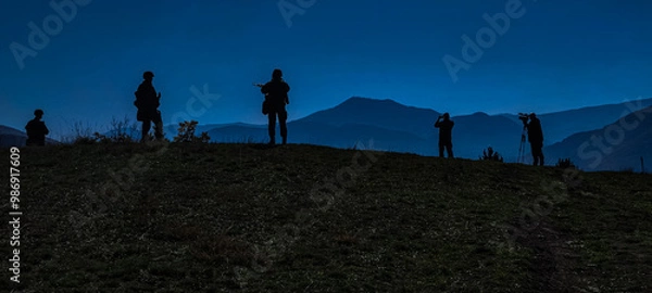 Obraz At night, on the top of a hill, there is an area where several people with silhouettes holding weapons and standing guard look into the camera