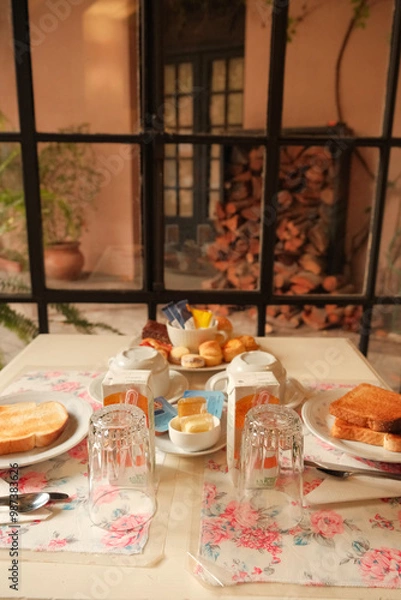 Fototapeta Pretty breakfast table setting in dining area by tall windows, in front of garden courtyard with firewood. Rustic cozy mood in hotel, hostel stay or guesthouse in Colonia del Sacramento, Uruguay