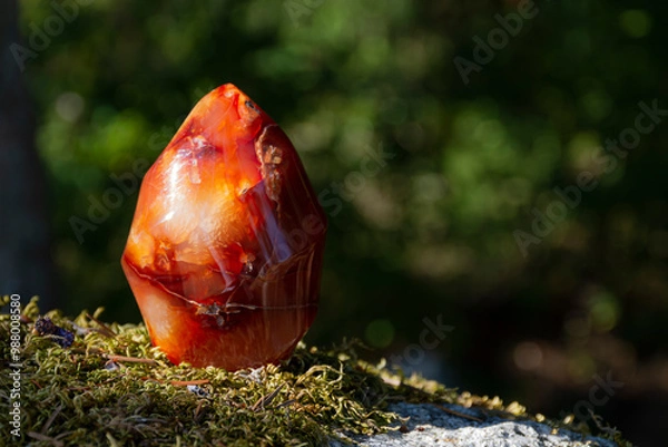 Fototapeta A close up image of a carnelian crystal flame tower glittering in the afternoon sunlight with dark green nature background. 