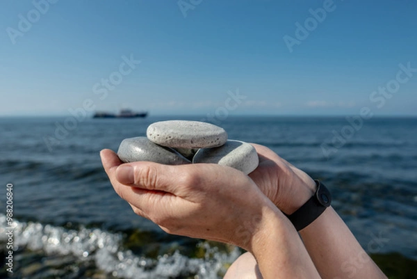Obraz Hands hold pieces of craft soap in the shape of stones against the background of the sea or Lake Baikal. Concept of the image of organic cosmetics against the background of nature