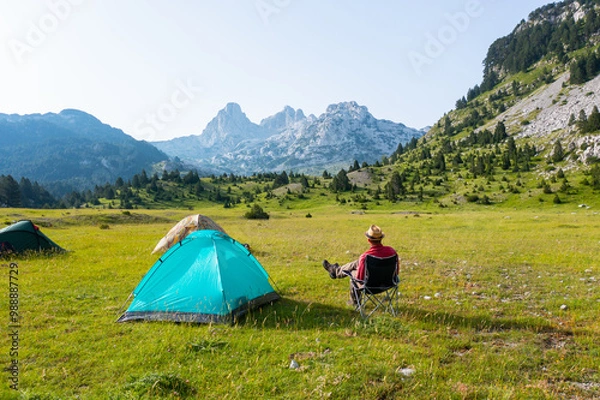 Fototapeta Hiker sitting next to a tent on a mountain in a beautiful valley with a view of rocky peaks in the background