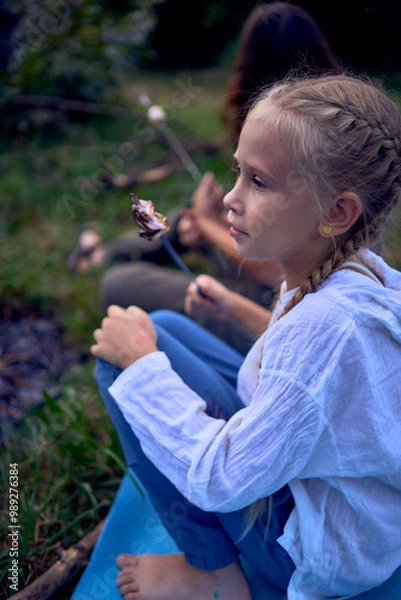 Fototapeta mother and her daughters  roasting and eating marshmallows on the campfire, camping vacation with children, mosquito bites on children's faces