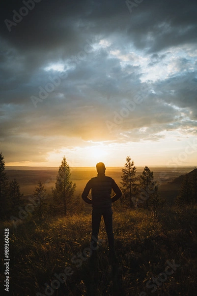 Fototapeta A man is standing at the very top of a picturesque hill, gazing at the beautiful colors of the sunset, completely enchanted by the view before him as the day transitions into night