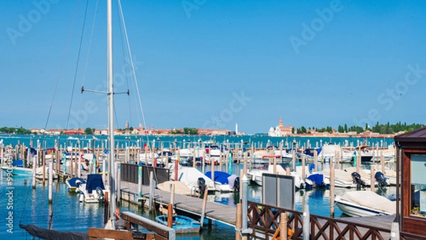 Fototapeta Petit port avec des bateaux à quai devant le quartier du Cannaregio à Venise dans la lagune avec la mer et un phare à l'arrière plan par une journée ensoleillée et un ciel bleu