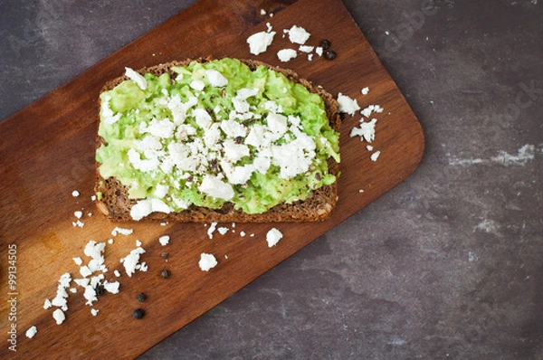 Obraz Healthy snack of dark rye bread topped with avocado and crumbled feta cheese served on a stone slate table top.