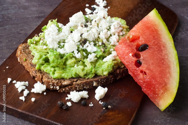 Obraz Healthy snack of dark rye bread topped with avocado and crumbled feta cheese served with a slice of fresh water melon.