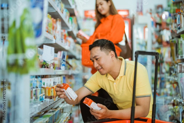 Obraz young Asian man squatting observing packaging while selecting goods in front of a shelf in supermarket