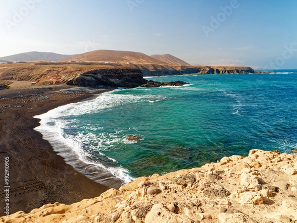 Fototapeta A beach with black volcanic sand in the town of Ajuy on the island of Fuerteventura is shown in this image. The rugged coastline meets clear turquoise waters, while barren hills rise in the background