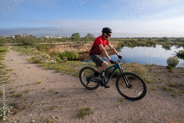 Fototapeta Mature Caucasian man riding a bicycle on a trail along a lake with trees and shrubs and city building in the background under hazy skies, Odessa Mountain Bike Park, Permian Basin, Texas