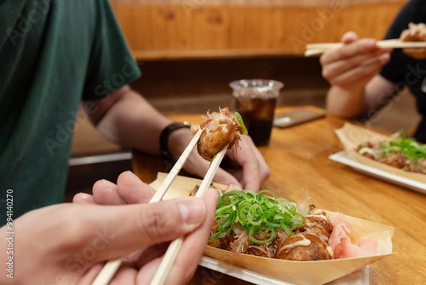Fototapeta A man is eating a takoyaki ball with his chopsticks in a bar with friends. A popular street food in Osaka, Japan