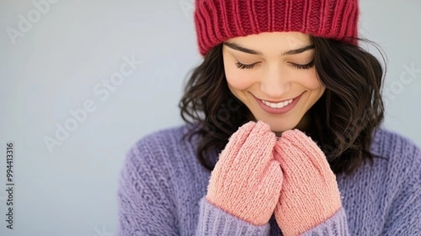 Obraz A young woman wearing a cozy sweater and hat smiles warmly while clasping her hands together
