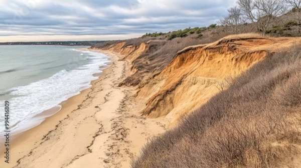 Fototapeta The view reveals sandy shores adjacent to towering cliffs that are gradually eroding, with waves softly lapping at the beach beneath a cloudy sky