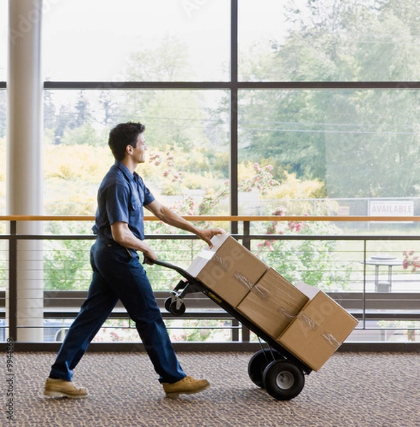 Fototapeta Delivery man in uniform pushing stack of boxes on dolly