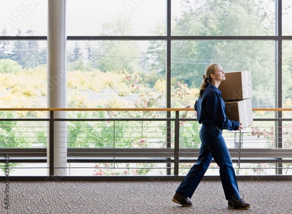 Obraz Side view of delivery woman in uniform carrying stack of boxes
