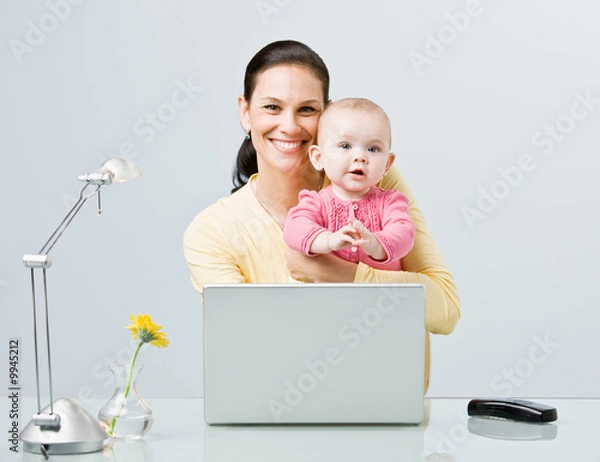 Fototapeta Working mother holding baby while typing on laptop at desk