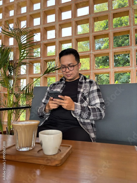 Fototapeta Man sitting in a cafe typing a message on his cell phone with coffee on the table