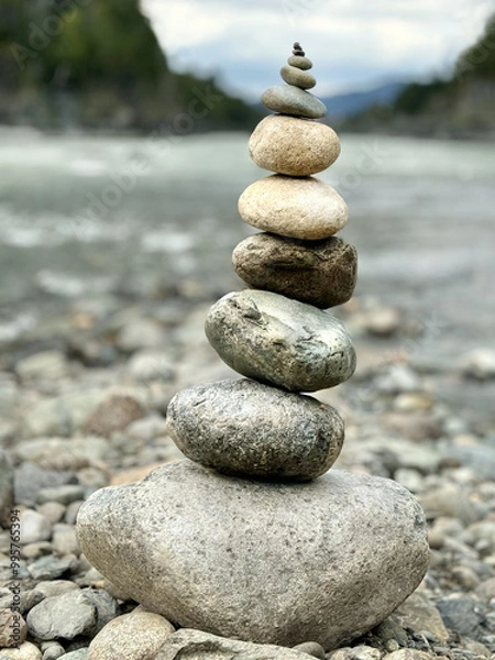 Obraz Round stones are stacked atop one another, set against the backdrop of a river and mountainous terrain, serving as a conceptual symbol of natural equilibrium.