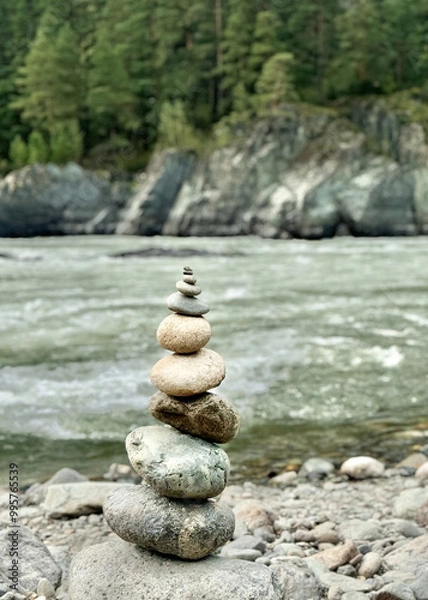 Obraz Smooth round stones are carefully balanced on top of each other, against the scenic view of a flowing river and rugged mountains, epitomizing the concept of natural balance.