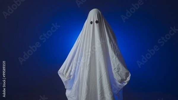 Fototapeta Funny Halloween ghost in white sheet makes scary gestures on background with blue spotlight in studio. Happy Halloween spooky concept.