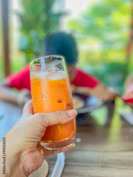 Obraz Closeup of glass of vibrant orange thai tea iced drink in a women hand with blurry backgrouhnd , relaxed atmosphere in a cafe or restaurant, enjoy a moment of refreshment., vertical color image.