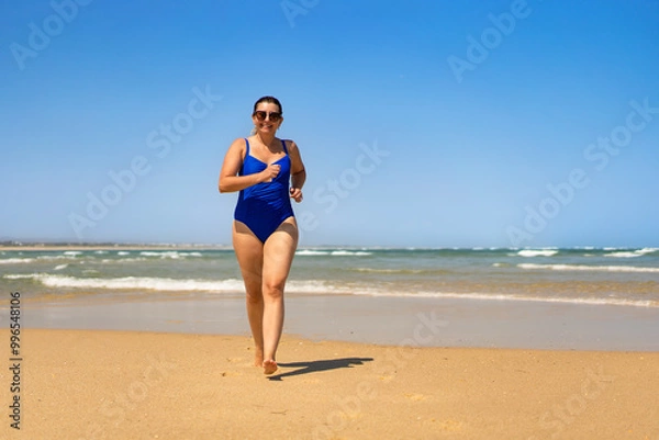 Fototapeta Beautiful middle-aged woman in one-piece blue swimsuit running on sandy beach in summertime. physical activity on beach. Front view