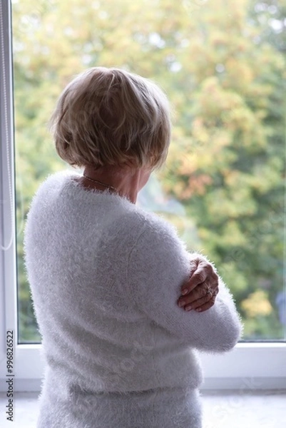 Obraz A lonely elderly woman standing at the window and waiting