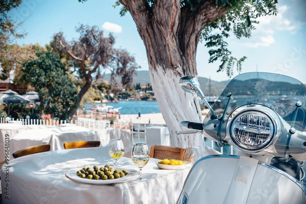 Obraz An ideal Greek setup by the sea in Neos Marmaras, parked scooter in the shadow of the tree, little table with Greek cuisine overlooking the port