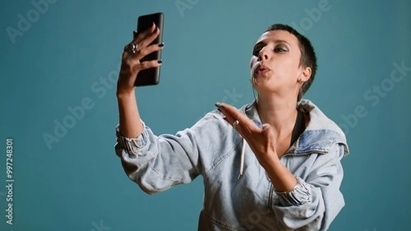 Fototapeta Silly stylish girl filming a video on her phone while she dances, showing funny groovy dance moves against blue background. Young gangster woman fooling around in studio. Camera A.