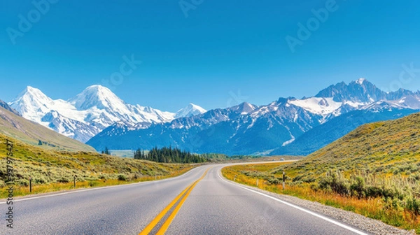 Fototapeta Scenic highway winding through lush green meadows with stunning snow-capped mountains under a clear blue sky