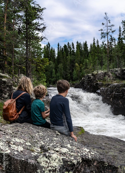 Fototapeta A mother and her two sons sit on a rock, captivated by the powerful Sognstupet Waterfall in Idre Dalarna surrounded by green trees and rushing water in the wilderness of Sweden