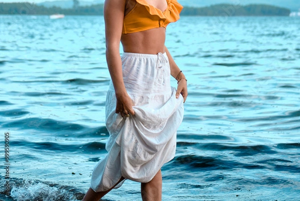 Fototapeta Una joven camina por la orilla del mar con la falda recogida para no mojarse.
