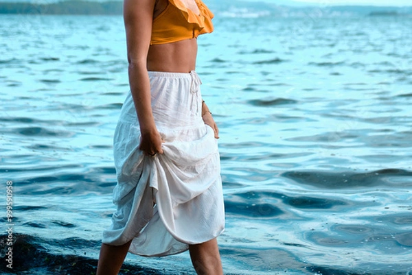 Fototapeta Vista lateral de una joven con una falda blanca de algodón recogida con las manos mientras pasea por la playa.