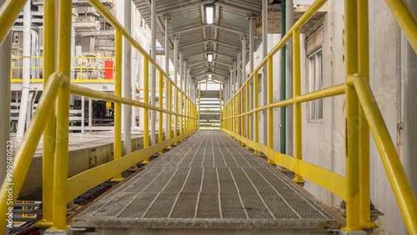Obraz Yellow Handrail Walkway in Industrial Setting. A yellow handrail walkway leads into a industrial area.  The walkway is made of metal and has a grid pattern.