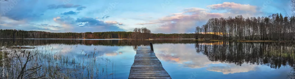 Fototapeta Krajobraz panoramiczny. Drewniane molo nad jeziorem o zachodzie słońca, odbicie chmur w wodzie.